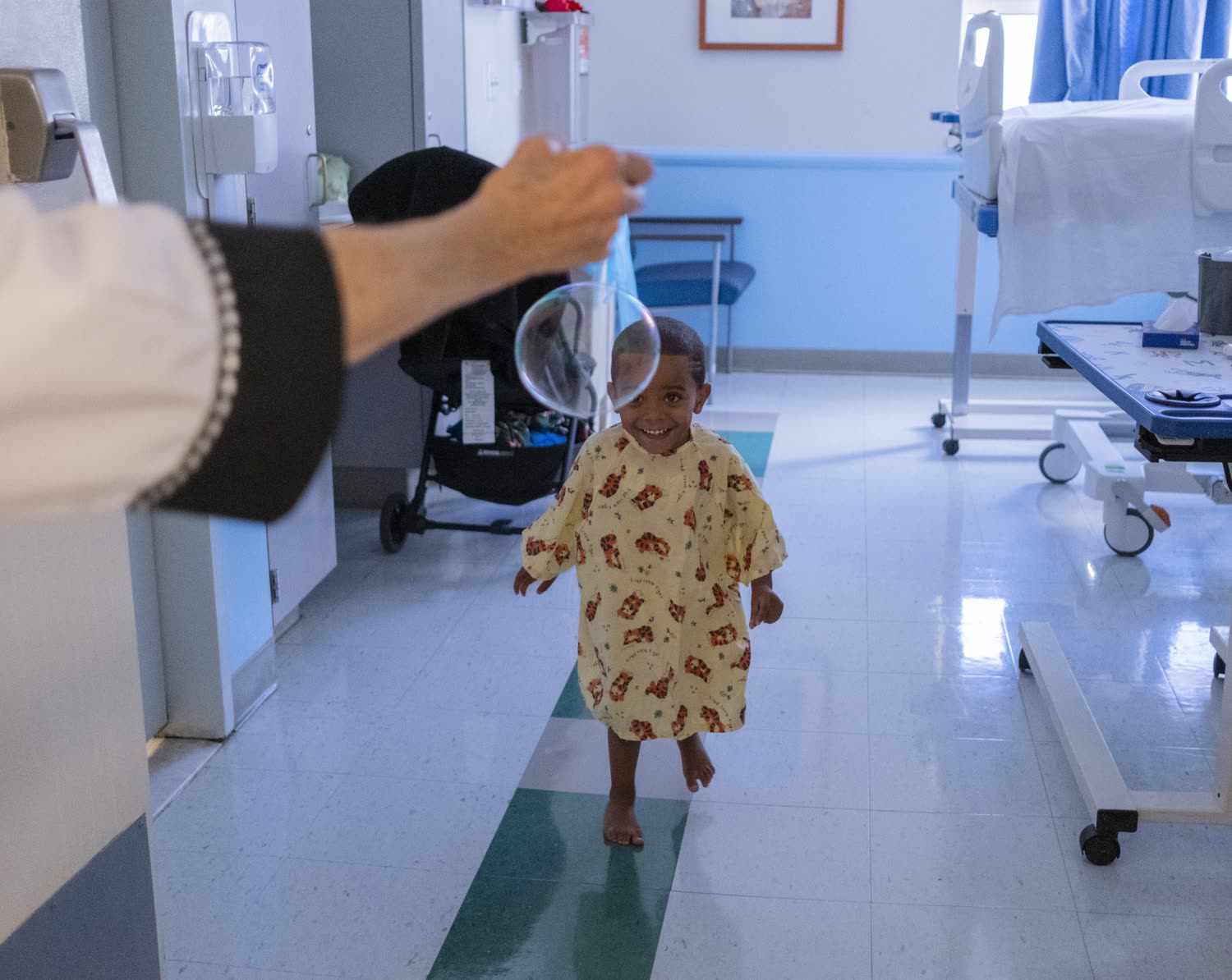 A small child in a hospital gown reaches for a bubble blown by a Red Nose Doc in a ward corridor.